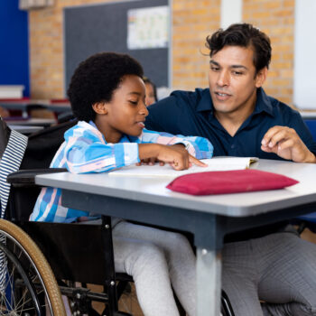Biracial male teacher helping schoolboy sitting in wheelchair in classroom at elementary school. Disability, education, childhood, development, learning and elementary school, unaltered.